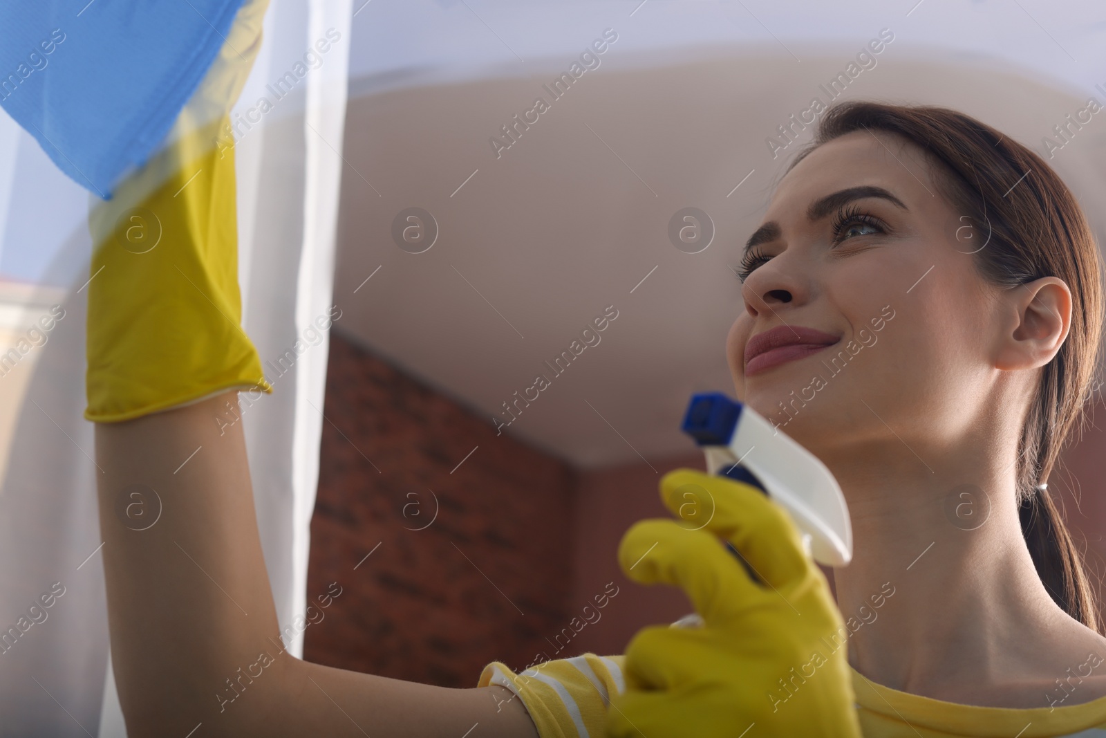 Photo of Young woman cleaning window glass with rag and detergent at home, view from outside