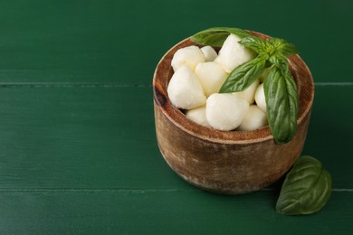 Photo of Tasty mozzarella balls and basil leaves in bowl on green wooden table. Space for text