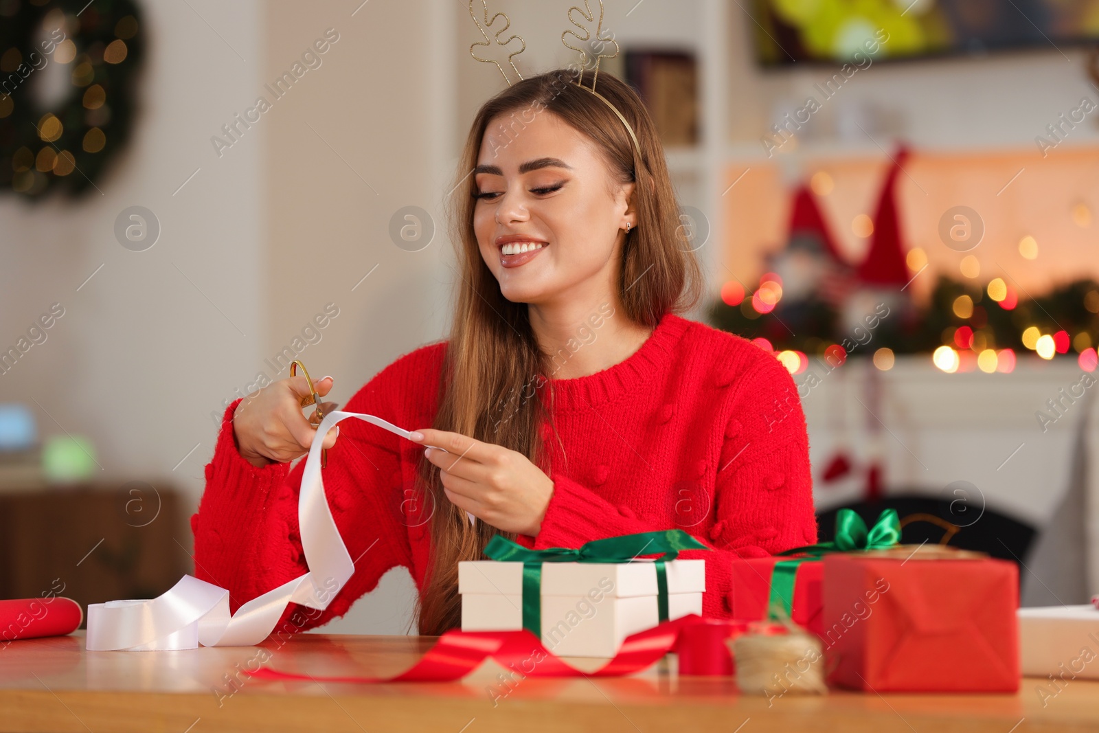 Photo of Beautiful young woman in deer headband cutting ribbon at table in room. Decorating Christmas gift