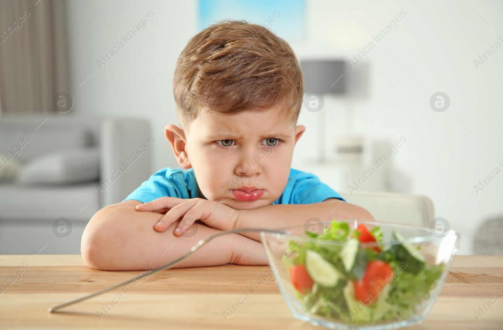 Photo of Stroppy little boy with bowl of vegetable salad at table in room