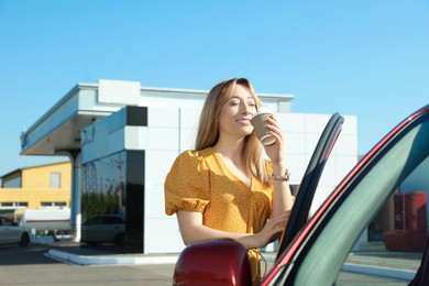 Beautiful young woman with coffee opening car door at gas station