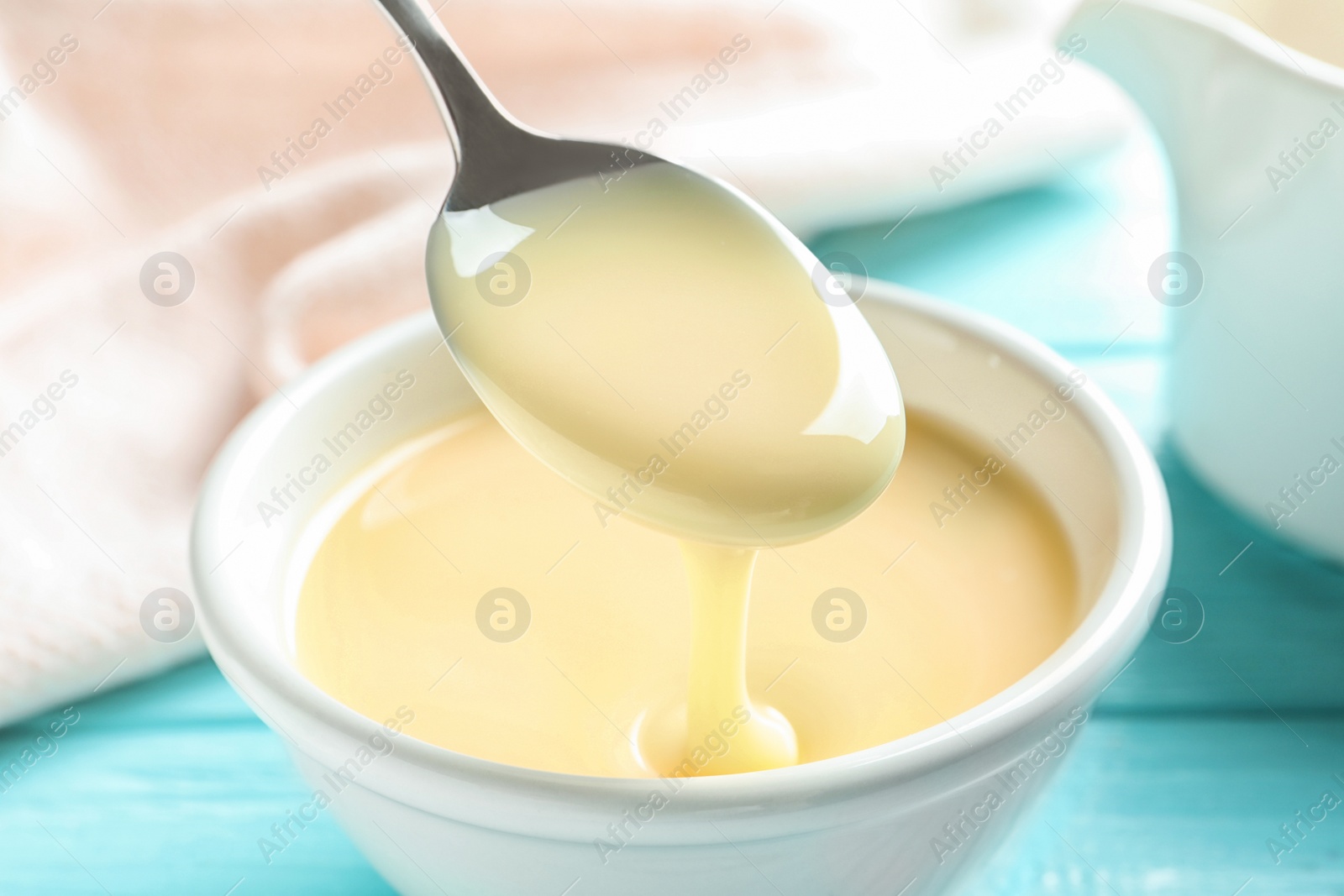 Photo of Spoon of pouring condensed milk over bowl on table, closeup. Dairy products