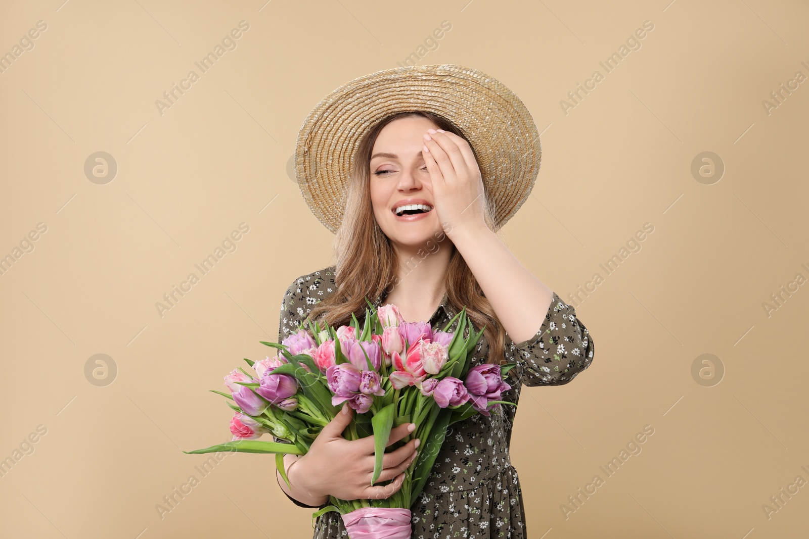 Photo of Happy young woman in straw hat holding bouquet of beautiful tulips on beige background