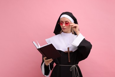 Woman in nun habit and sunglasses reading Bible against pink background