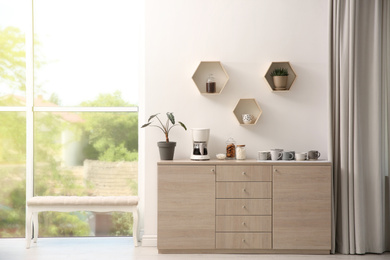 Photo of Modern coffeemaker and cups on commode near light wall indoors