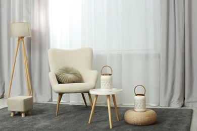 Photo of Comfortable armchair, white table and stylish lanterns near elegant curtains in light room. Interior design