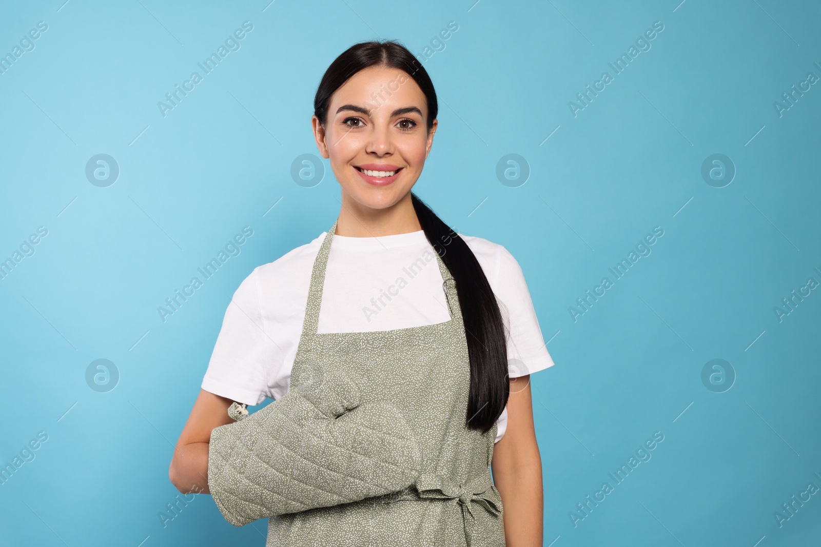 Photo of Young woman in grey apron and oven glove on light blue background, space for text