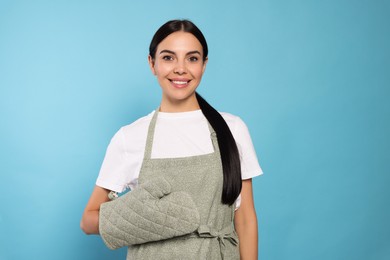 Young woman in grey apron and oven glove on light blue background, space for text