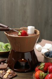Fondue pot with melted chocolate, different fresh berries, kiwi, marshmallows and forks on wooden table