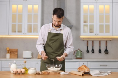 Making bread. Man putting dry yeast into bowl with flour at wooden table in kitchen