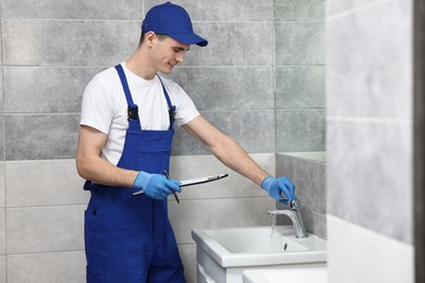 Smiling plumber with clipboard examining faucet in bathroom