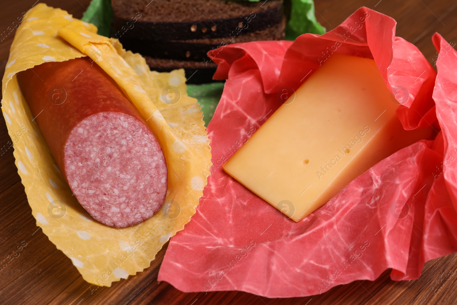 Photo of Different products in beeswax food wraps on wooden table, closeup