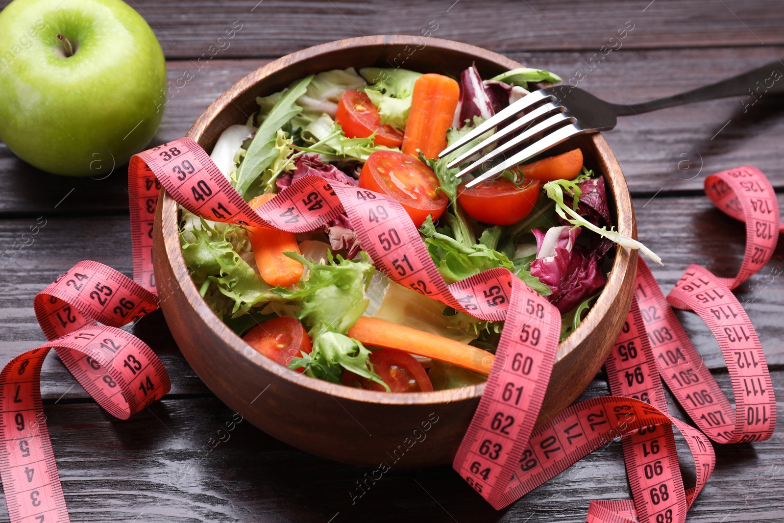 Photo of Bowl with fresh vegetable salad, fork, apple and measuring tape on wooden table, closeup. Healthy diet concept