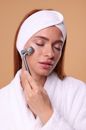 Young woman massaging her face with metal roller on pale orange background