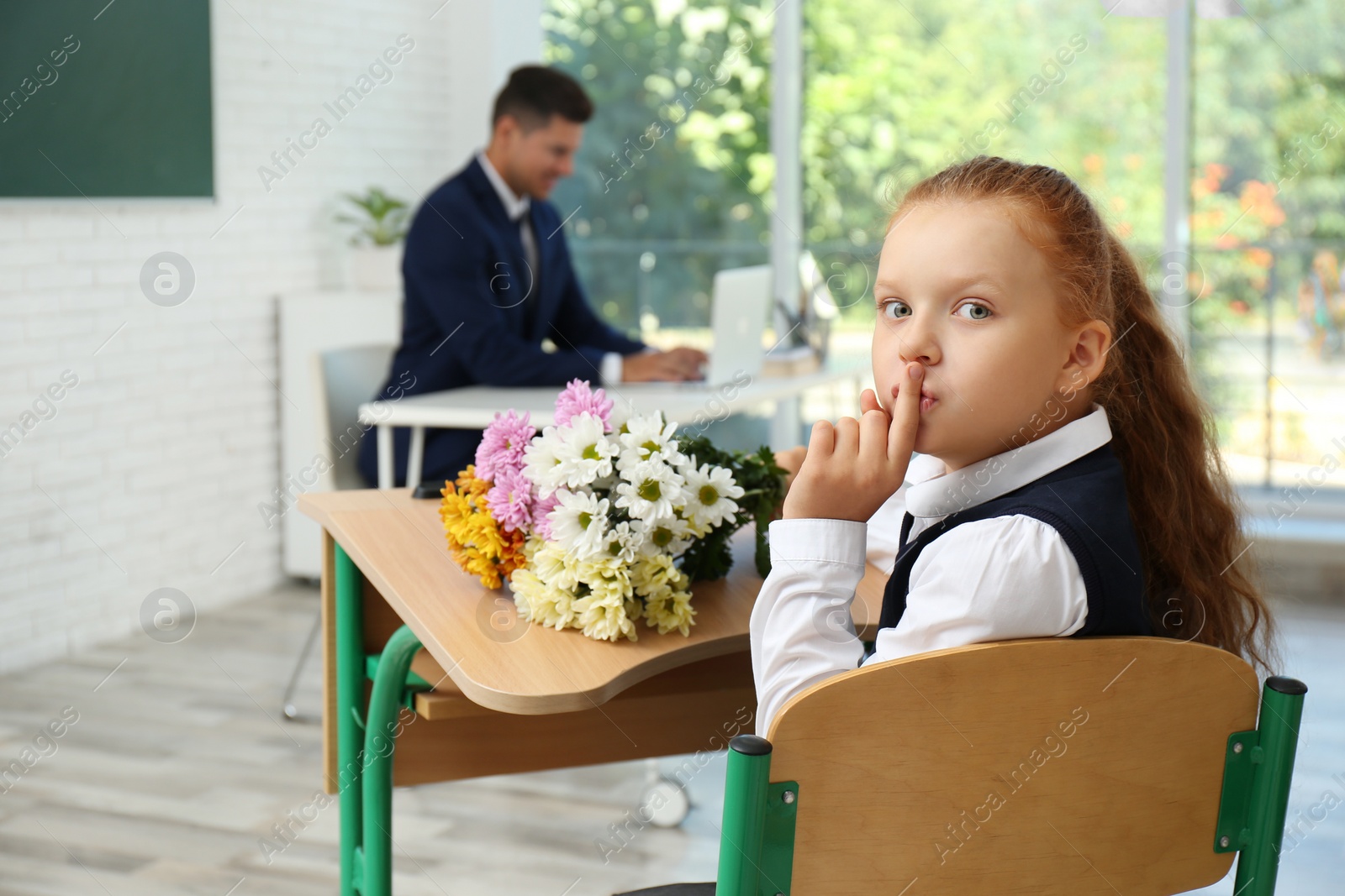 Photo of Happy schoolgirl with bouquet sitting at desk in classroom. Teacher's day