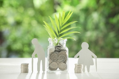 Pension savings. Figures of elderly people, jar with coins and twig on white table against blurred green background