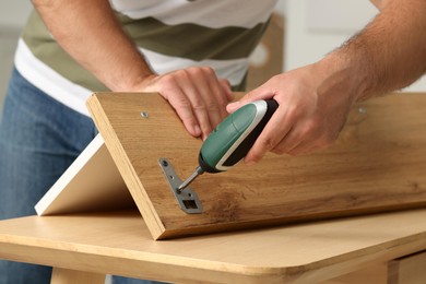 Photo of Man with electric screwdriver assembling furniture at table indoors, closeup