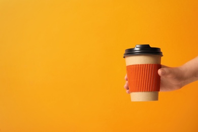 Woman holding takeaway paper coffee cup with cardboard sleeve on orange background, closeup. Space for text