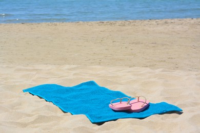Photo of Soft light blue beach towel and pink flip flops on sand near sea. Space for text