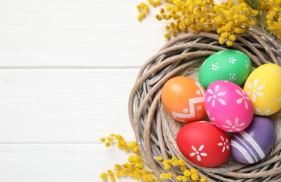 Photo of Colorful Easter eggs in decorative nest and mimosa flowers on white wooden background, flat lay. Space for text