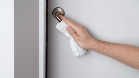 Photo of Man cleaning doorknob with wet wipe indoors, closeup. Protective measures