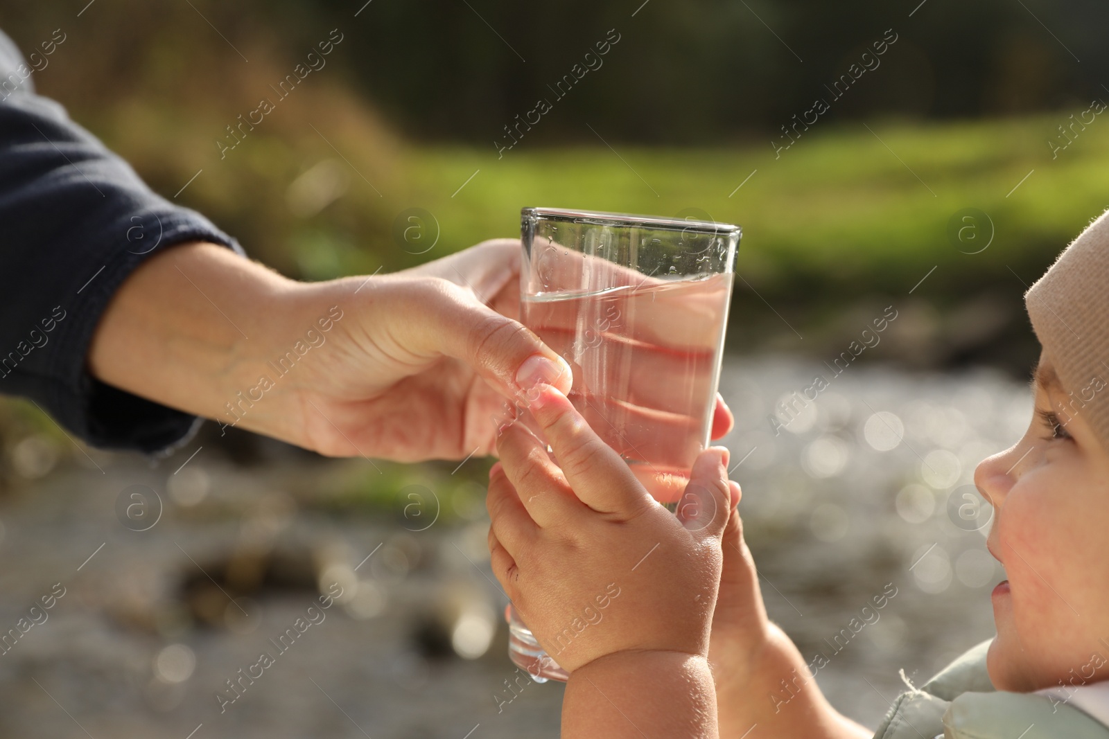 Photo of Mother giving her daughter glass of fresh water near stream on sunny day, closeup