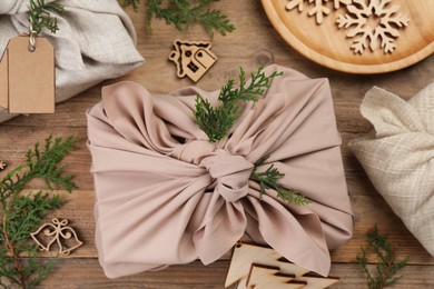 Photo of Furoshiki technique. Gift packed in fabric, thuja branches and festive decoration on wooden table, flat lay