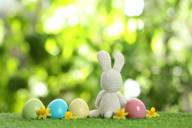 Cute Easter bunny toy and dyed eggs on green grass against blurred background, space for text