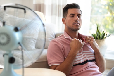 Photo of Man enjoying air flow from fan in living room. Summer heat