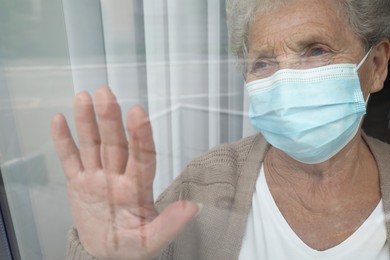 Photo of Senior woman in protective mask looking out of window at nursing home, view from outside
