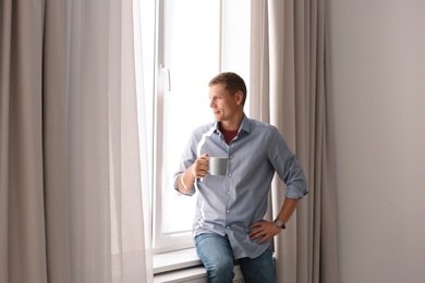 Photo of Mature man with cup of hot drink near window with open curtains at home