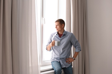 Mature man with cup of hot drink near window with open curtains at home