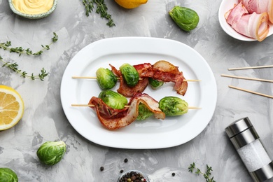 Photo of Skewers with Brussels sprouts and bacon in plate on grey table, flat lay