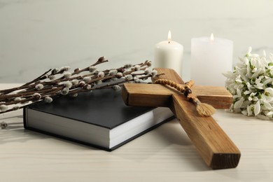 Burning church candles, wooden cross, rosary beads, Bible, willow branches and snowdrops on white table