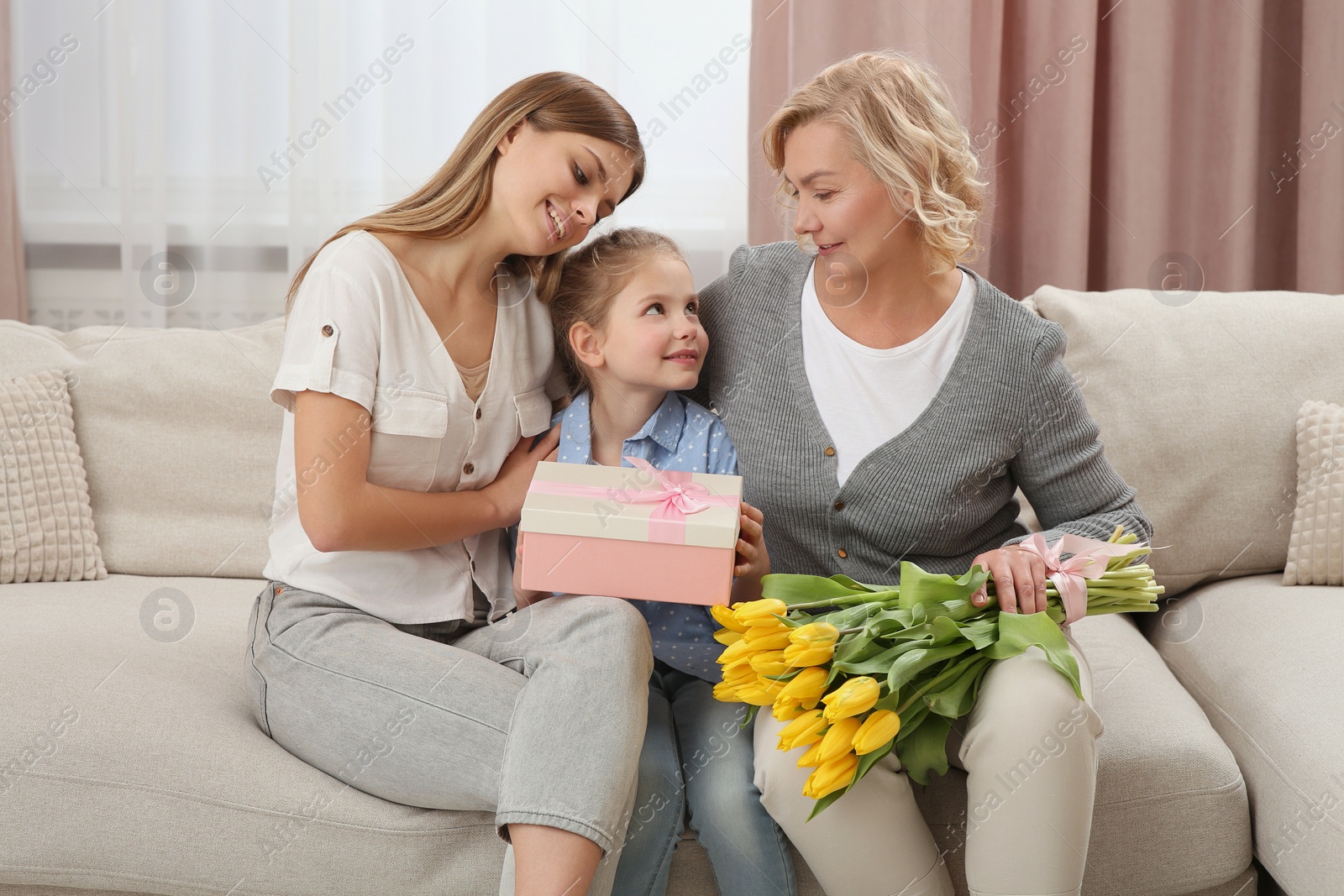 Photo of Little girl congratulating her mom and granny with flowers and gift at home. Happy Mother's Day