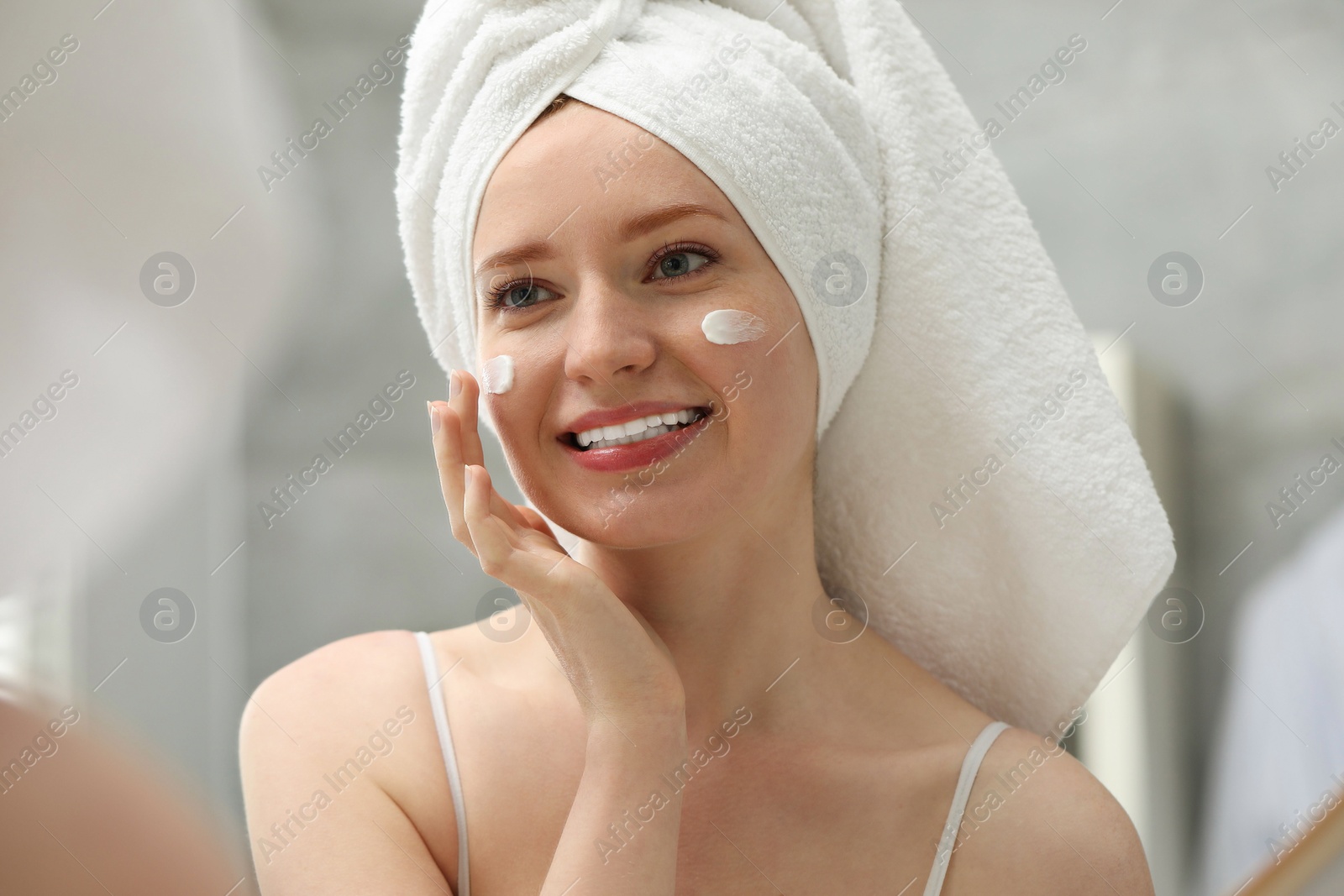 Photo of Smiling woman with freckles applying cream onto her face near mirror in bathroom