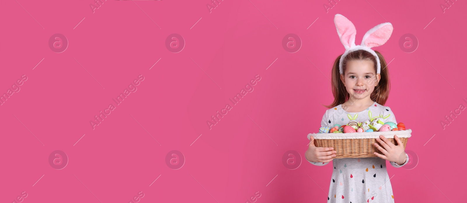 Photo of Adorable little girl with bunny ears holding wicker basket full of Easter eggs on pink background. Space for text