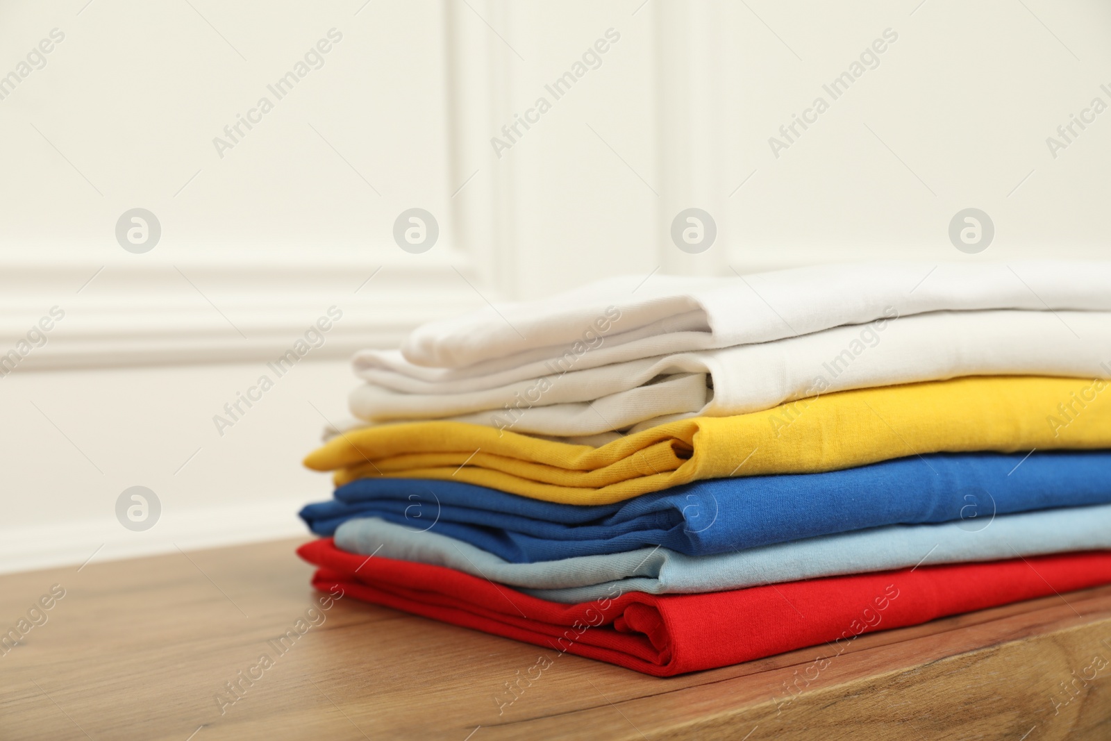 Photo of Sorting and organizing. Stack of different folded clothes on wooden table near white wall, closeup with space for text