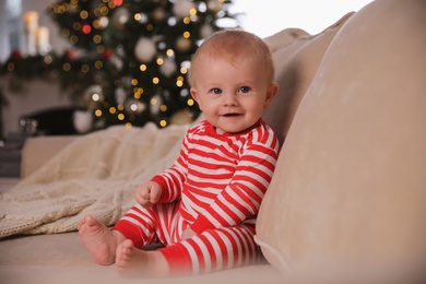 Cute baby in bright Christmas pajamas sitting on sofa at home