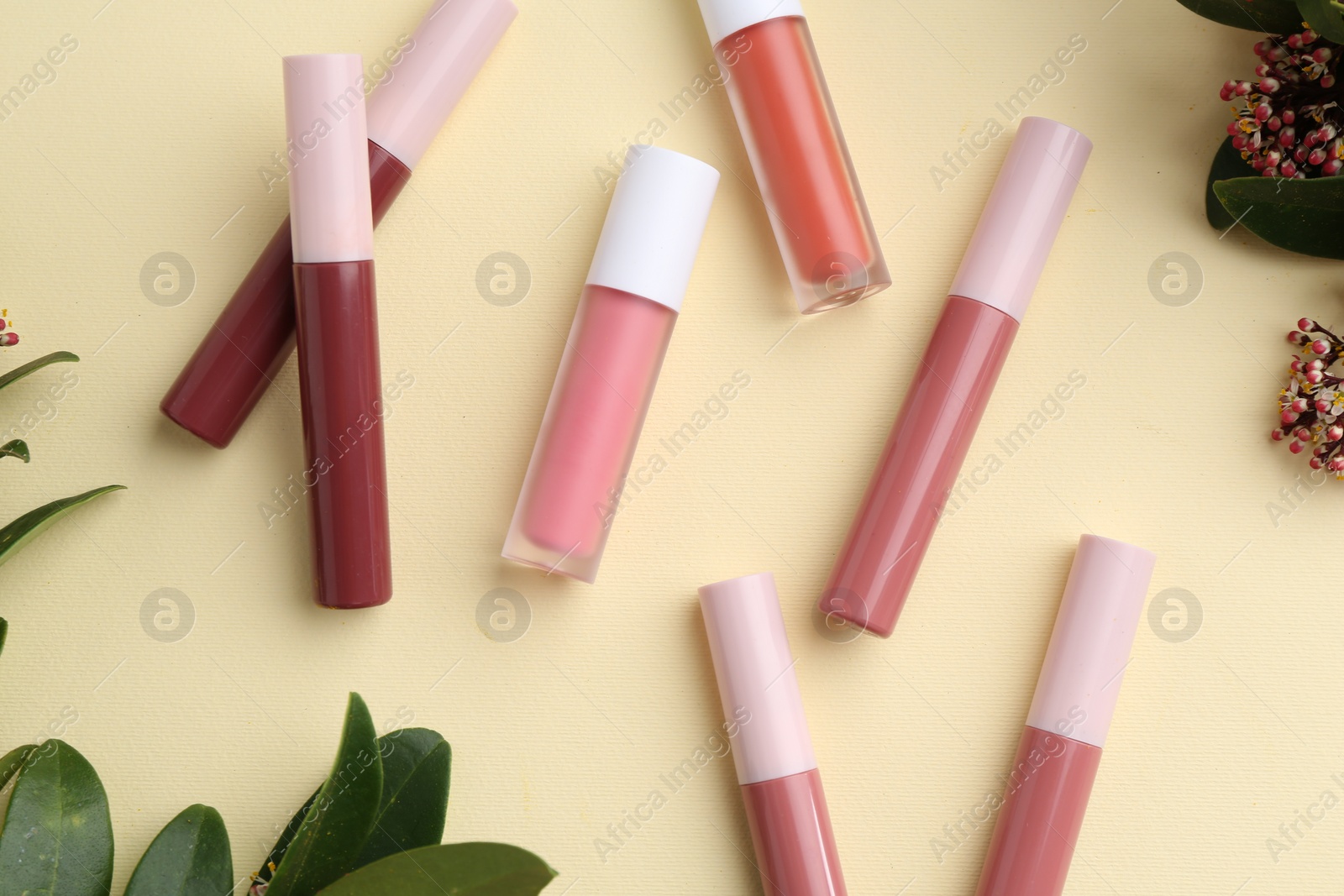 Photo of Different lip glosses, green leaves and flowers on pale yellow background, flat lay