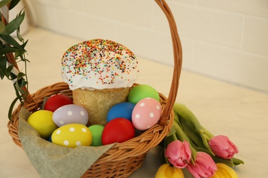 Photo of Basket with traditional Easter cake, dyed eggs and flowers on white table