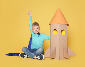 Little child in cape playing with rocket made of cardboard box on yellow background