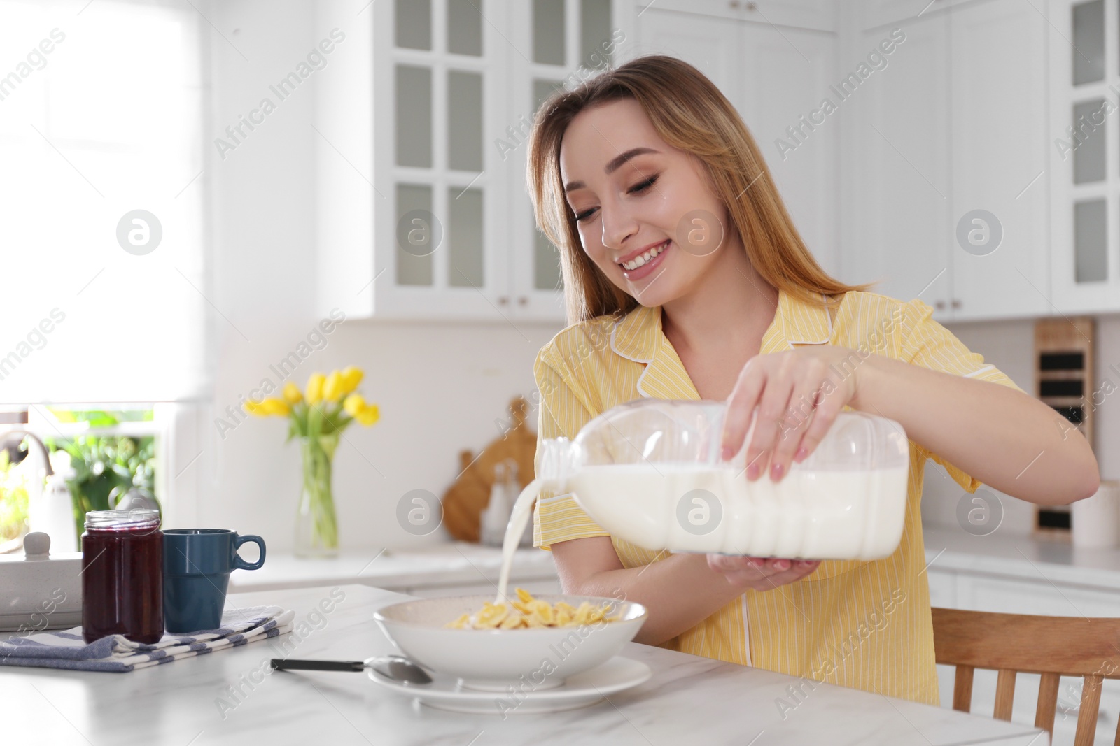 Photo of Young woman pouring milk from gallon bottle into plate with breakfast cereal at white marble table in kitchen