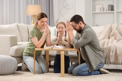 Photo of Girl and her godparents praying over Bible together at table indoors