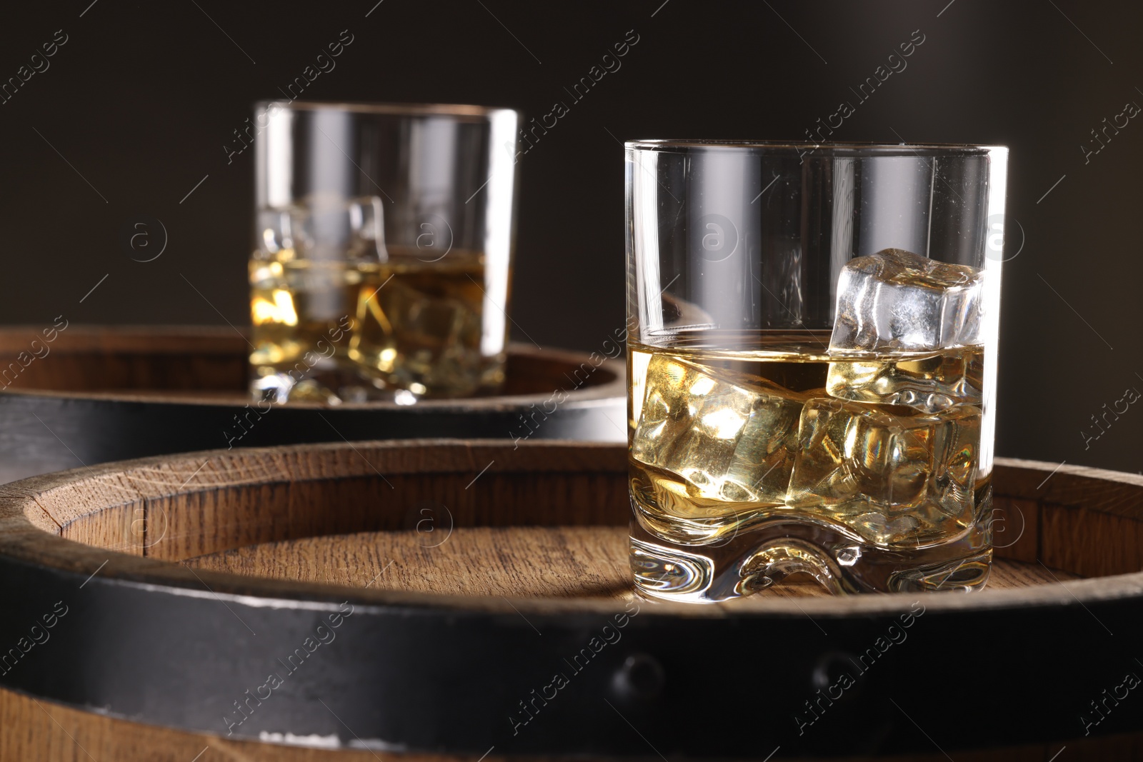 Photo of Whiskey with ice cubes in glasses on wooden barrels against dark background, closeup