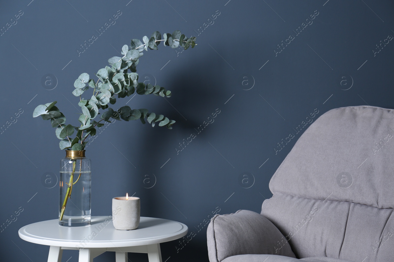 Photo of Beautiful eucalyptus branches and burning candle on white table near blue wall. Interior element