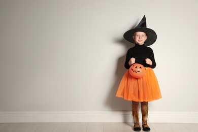 Photo of Cute little girl with pumpkin candy bucket wearing Halloween costume near light wall. Space for text