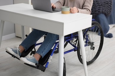 Photo of Woman in wheelchair with cup of drink using laptop indoors, closeup
