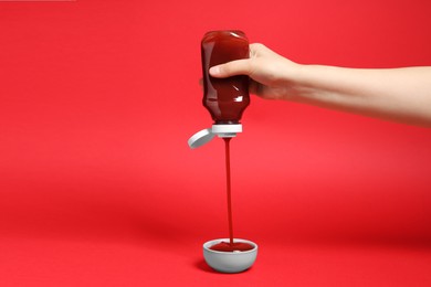 Photo of Woman pouring tasty ketchup from bottle into bowl on red background, closeup