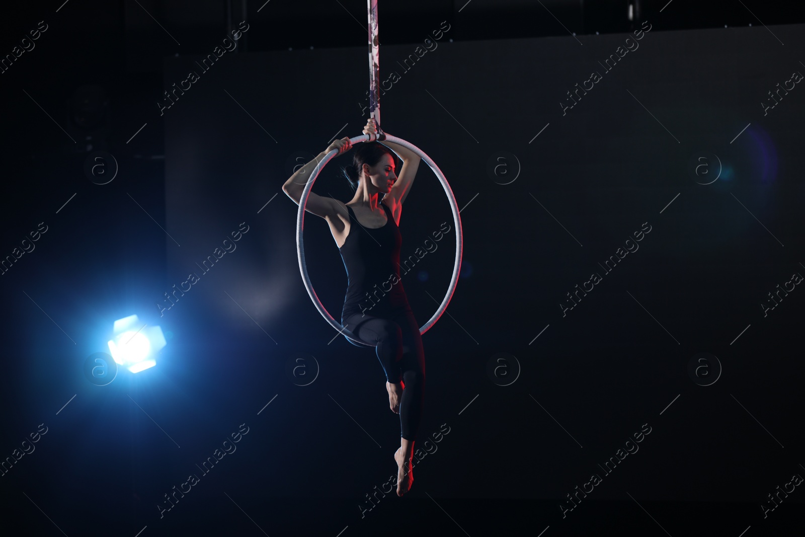 Photo of Young woman performing acrobatic element on aerial ring against dark background